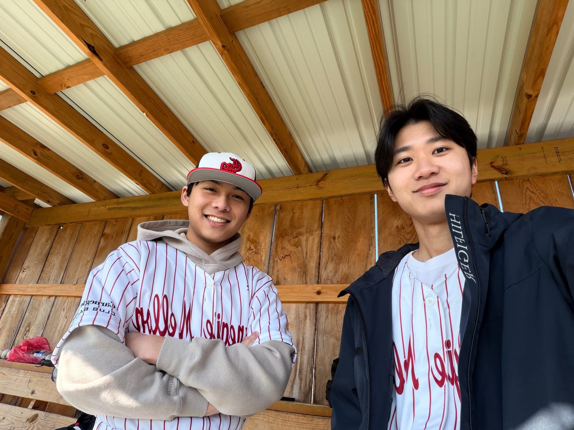 Two teammates in red-pinstriped Carnegie Mellon baseball jerseys smiling in a wooden dugout