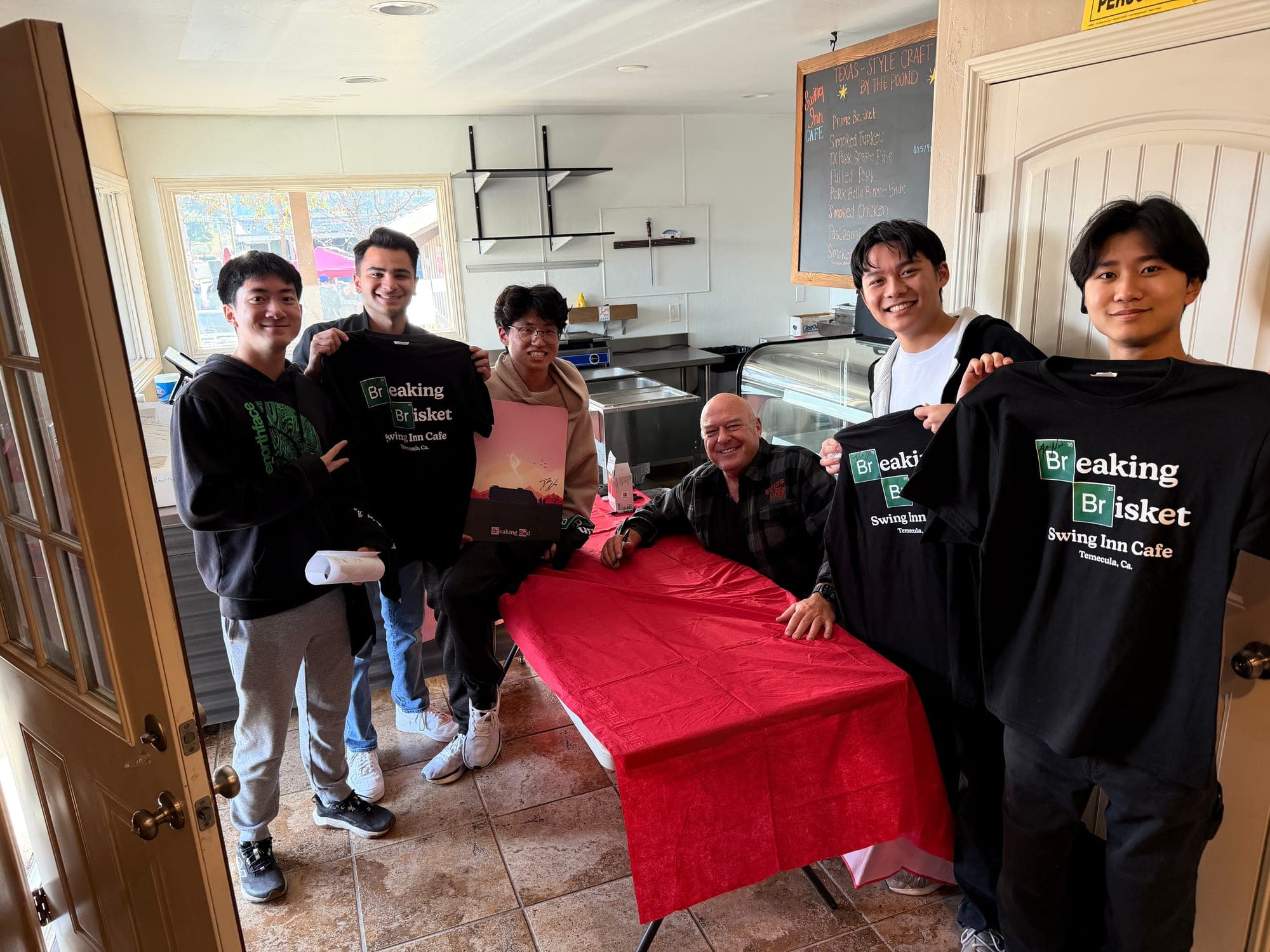 Friends holding up Breaking Brisket T-shirts around a signing table at Swing Inn Cafe with the actor who played Hank Schrader