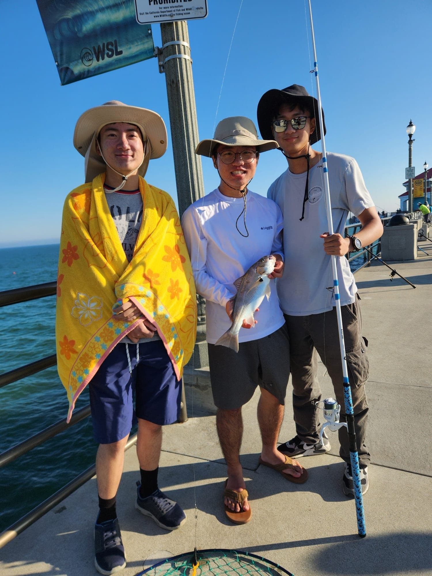 Three friends in sun hats on a pier, one holding up a fish they caught