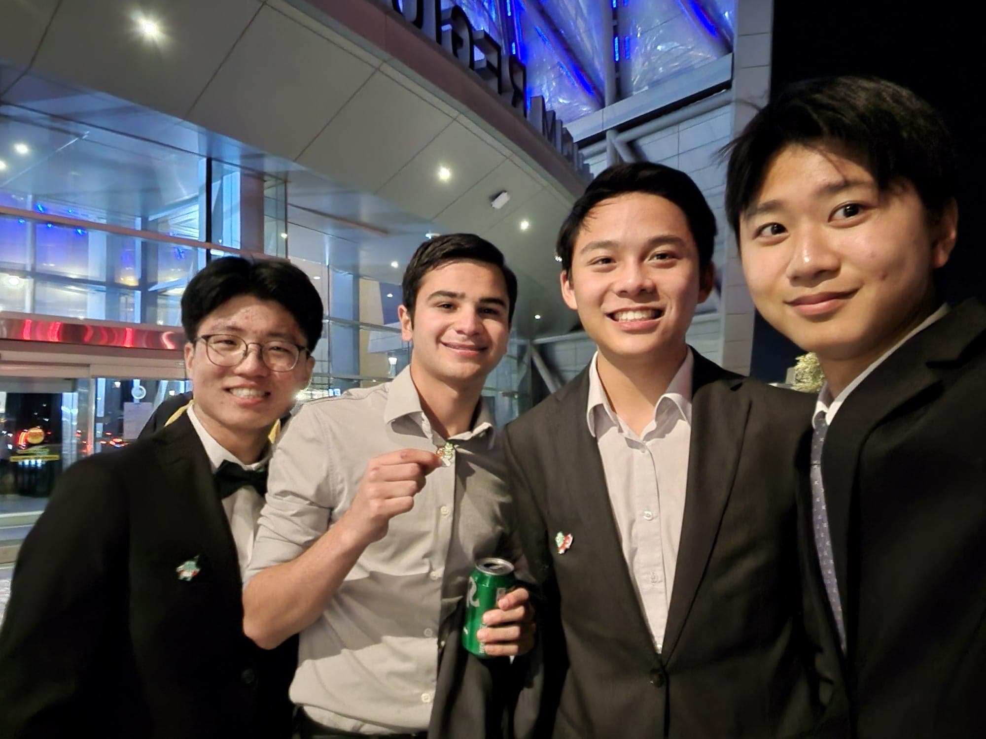 Four friends in suits smiling for a selfie outside a lit-up arena at night