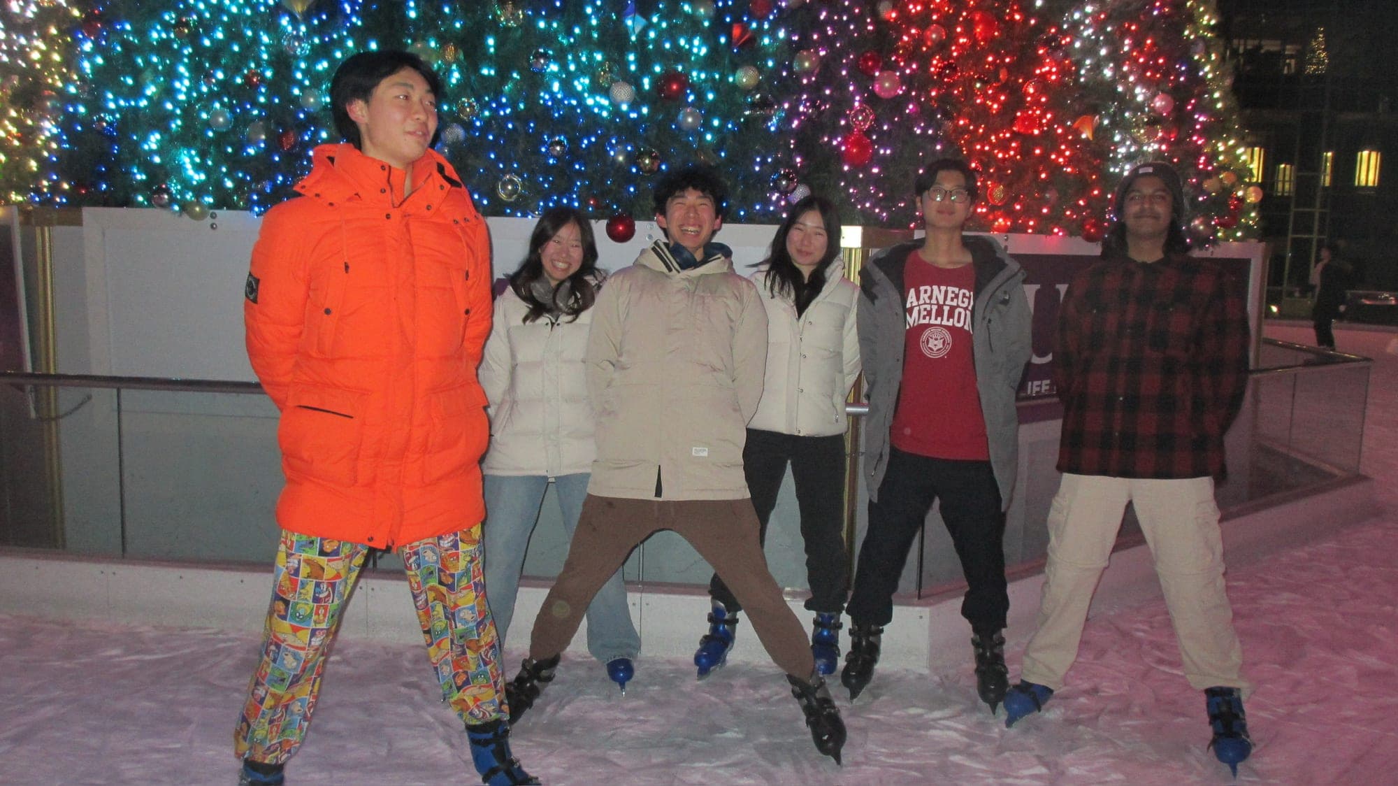 Six friends in winter coats on ice skates, posing on a rink in front of a big lit-up Christmas tree at night