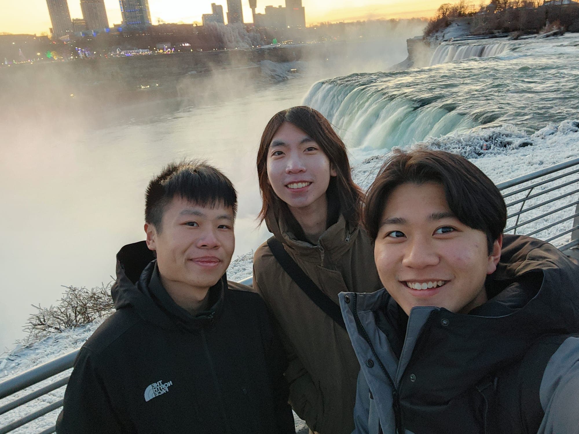 Three friends in winter jackets taking a selfie in front of a misty waterfall and frosted railings at sunset