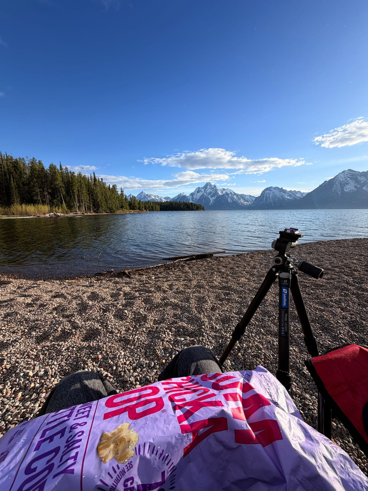 A tripod on a pebble beach by a lake, with snow-capped mountains across the water and a bag of kettle corn in the foreground