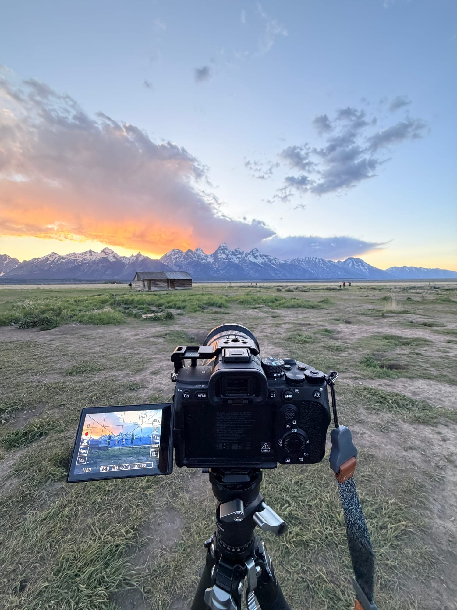 A camera on a tripod framing an old wooden barn and jagged snowy peaks under an orange sunset
